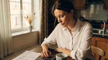 Une femme assise à sa table de cuisine utilise une calculatrice devant une facture pour estimer le montant du chèque énergie de son foyer